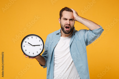 Exhausted young man in casual blue shirt posing isolated on yellow orange wall background, studio portrait. People emotions lifestyle concept. Mock up copy space. Holding clock, putting hand on head.