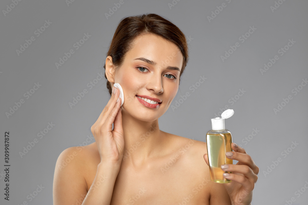beauty, skin care and people concept - smiling young woman with toner or cleanser and cotton pad cleansing face over grey background beauty, skin care and people concept - smiling young woman with toner or cleanser and cotton pad cleansing face over grey background