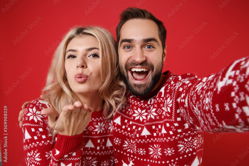 Close up of merry young couple guy girl in Christmas sweaters posing isolated on red background. Happy New Year 2020 celebration party concept. Mock up copy space. Doing selfie shot on mobile phone. Close up of merry young couple guy girl in Christmas sweaters posing isolated on red background. Happy New Year 2020 celebration party concept. Mock up copy space. Doing selfie shot on mobile phone.
