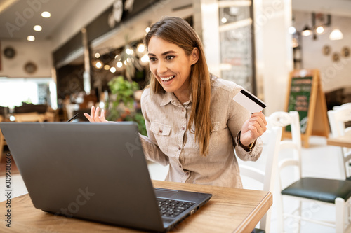 Beautiful young caucasian woman with brown hair, smiling while holding her credit card in a coffee shop. She's looking at her laptop, pleasantly surprised with online shopping.