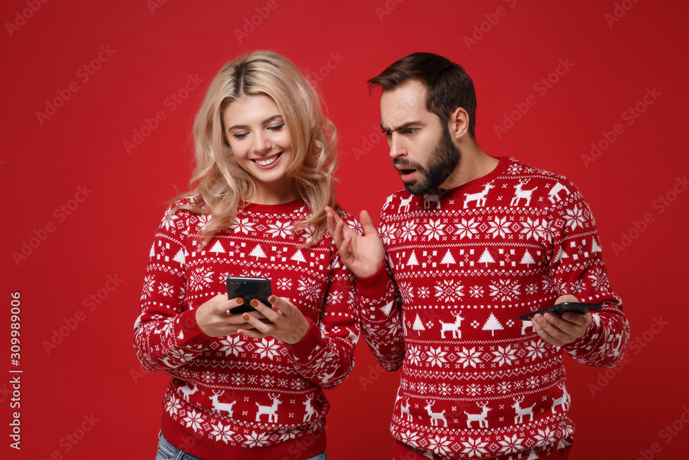 Shocked young couple guy girl in Christmas knitted sweaters posing isolated on red background. New Year 2020 celebration holiday concept. Mock up copy space. Using mobile phone typing sms message. Shocked young couple guy girl in Christmas knitted sweaters posing isolated on red background. New Year 2020 celebration holiday concept. Mock up copy space. Using mobile phone typing sms message.
