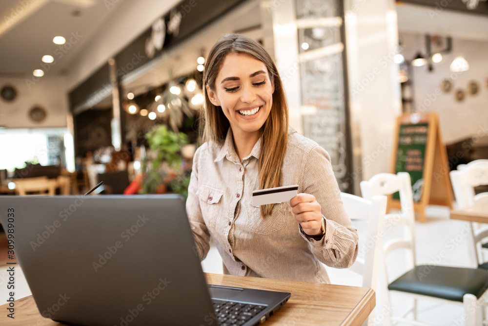 Beautiful young caucasian woman smiling looking at credit card she's holding and checking her account number. She's relaxed and cheerful while online shopping in a coffee shop. Beautiful young caucasian woman smiling looking at credit card she's holding and checking her account number. She's relaxed and cheerful while online shopping in a coffee shop.