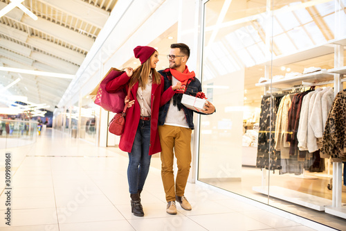 Joyful young caucasian couple going Christmas shopping, walking by the shop window embraced. They are embracing and looking each other and carrying shopping bags and gift in shopping mall.