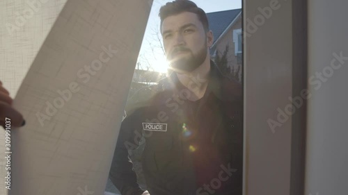 Portrait of young Caucasian police officer standing behind glass door and looking inside. Female hand closing curtain. Law enforcement, police patrol. photo