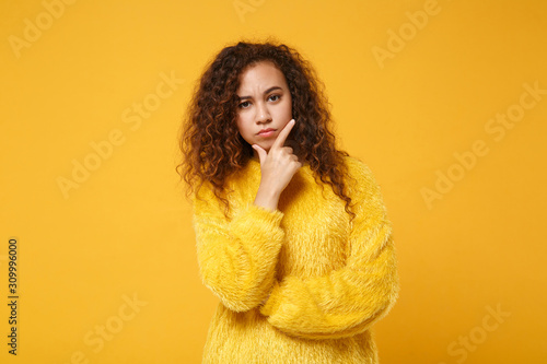 Pensive young african american girl in fur sweater posing isolated on yellow orange background studio portrait. People sincere emotions lifestyle concept. Mock up copy space. Put hand prop up on chin. © ViDi Studio