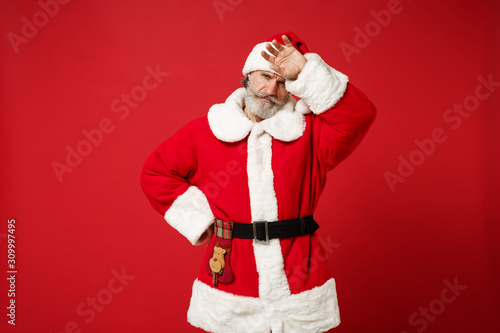 Tired elderly gray-haired mustache bearded Santa man in Christmas hat posing isolated on red wall background. Happy New Year 2020 celebration holiday concept. Mock up copy space. Putting hand on head. photo