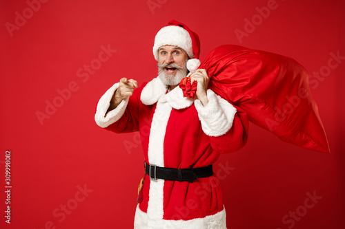 Elderly gray-haired mustache bearded Santa man in Christmas hat isolated on red background. New Year 2020 celebration holiday concept. Mock up copy space. Hold bag with gifts, point finger on camera.