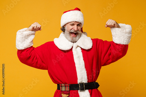 Funny elderly gray-haired mustache bearded Santa man in Christmas hat posing isolated on yellow wall background. Happy New Year 2020 celebration concept. Mock up copy space. Showing biceps, muscles.