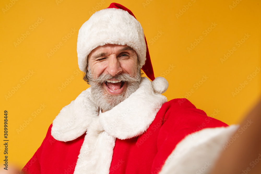Close up of elderly gray-haired bearded mustache Santa man in Christmas hat posing isolated on yellow background. New Year 2020 holiday concept. Mock up copy space. Doing selfie shot on mobile phone. Close up of elderly gray-haired bearded mustache Santa man in Christmas hat posing isolated on yellow background. New Year 2020 holiday concept. Mock up copy space. Doing selfie shot on mobile phone.