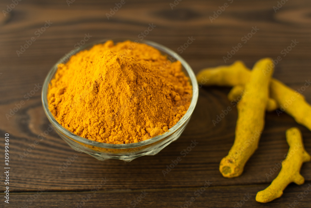 Blurred turmeric powder in glass bowl and turmeric roots on wooden background. Blurred turmeric powder in glass bowl and turmeric roots on wooden background.