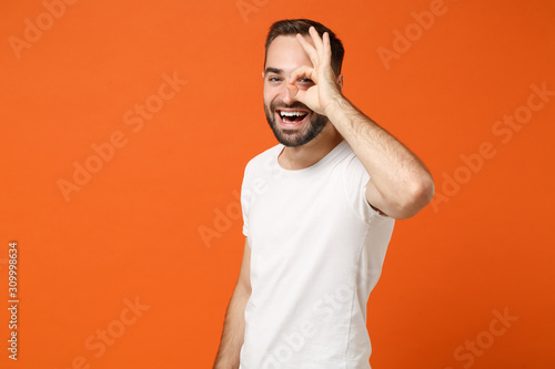 Funny young man in casual white t-shirt posing isolated on orange wall background, studio portrait. People lifestyle concept. Mock up copy space. Holding hand near eye imitating binoculars or glasses.