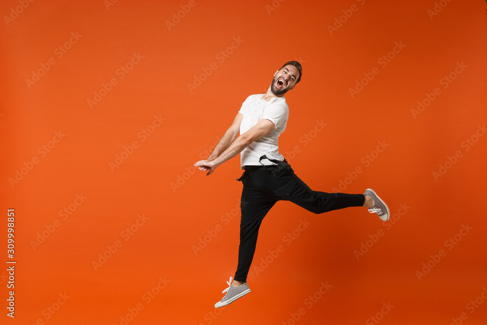 Cheerful crazy young man in casual white t-shirt posing isolated on bright orange wall background studio portrait. People lifestyle concept. Mock up copy space. Having fun, fooling around, jumping. Cheerful crazy young man in casual white t-shirt posing isolated on bright orange wall background studio portrait. People lifestyle concept. Mock up copy space. Having fun, fooling around, jumping.