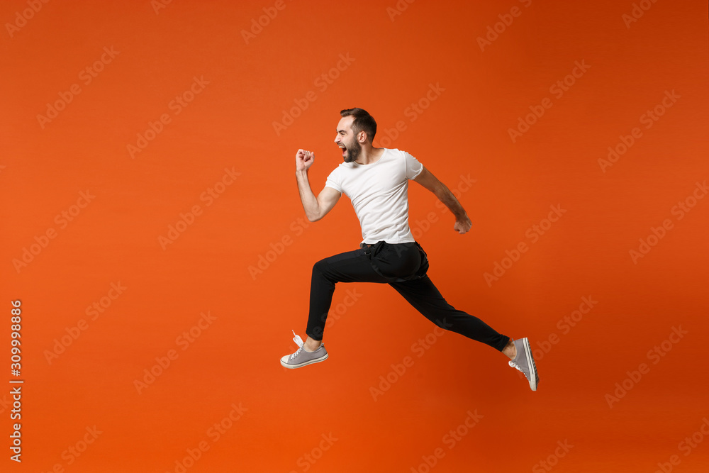 Side view of crazy young man in casual white t-shirt posing isolated on  bright orange wall background studio portrait. People lifestyle concept.  Mock up copy space. Having fun fooling around, jumping. Stock