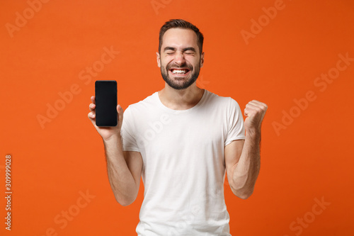 Happy young man in white t-shirt posing isolated on orange background. People lifestyle concept. Mock up copy space. Hold mobile phone with blank empty screen doing winner gesture keeping eyes closed.