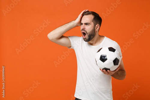 Shocked young man in casual white t-shirt posing isolated on orange background studio portrait. People sincere emotions lifestyle concept. Mock up copy space. Holding soccer ball putting hand on head.