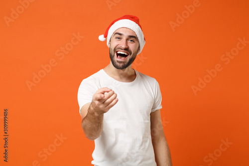 Cheerful young Santa man in white t-shirt, Christmas hat posing isolated on orange background. Happy New Year 2020 celebration holiday concept. Mock up copy space. Pointing index finger on camera.
