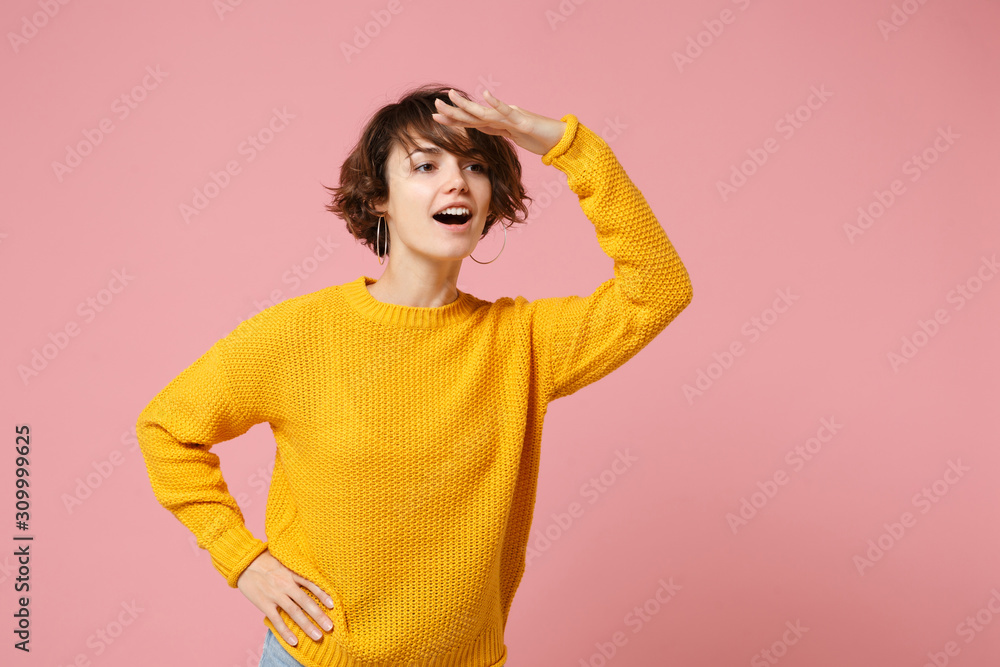 Obraz premium Curious young brunette woman girl in yellow sweater posing isolated on pastel pink background in studio. People lifestyle concept. Mock up copy space. Hold hand at forehead looking far away distance.
