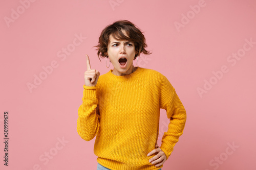 Displeased young brunette woman girl in yellow sweater posing isolated on pastel pink wall background studio portrait. People lifestyle concept. Mock up copy space. Swearing, pointing index finger up.