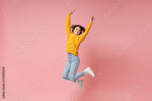 Smiling young brunette woman girl in yellow sweater posing isolated on pastel pink background in studio. People lifestyle concept. Mock up copy space. Having fun fooling around, rising hands, jumping.