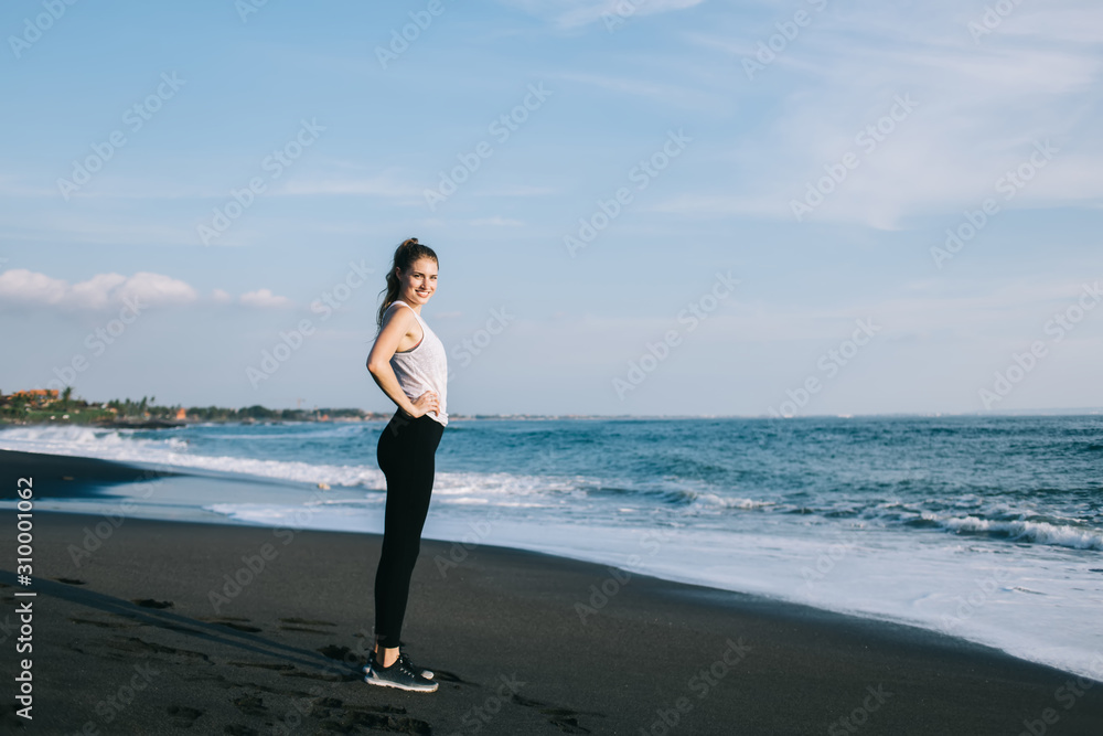 Young smiling brunette in sportswear with hands on waist against sky and sea