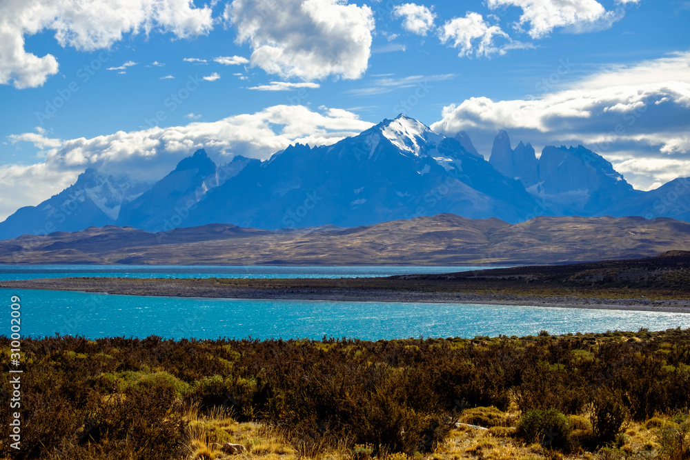 Under a blue, partially clouded sky, you can see the famous 'Torres del Paine'. These mountain peaks are the destination of a stunning trekking circuit. In the foreground lies Lake Sarmiento.