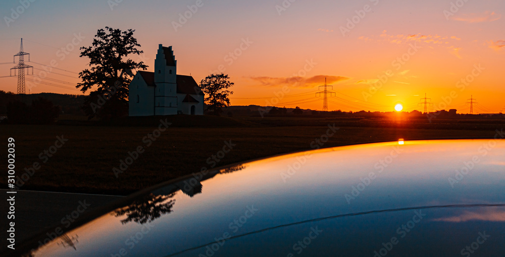 Beautiful sunset with a church and reflections on a car roof near Huett, Bavaria, Germany