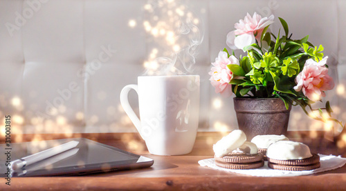 Wooden table, tray, white cup of coffee, flowers on the table, tablet. Sunlight, ray. Abstract bokeh light.