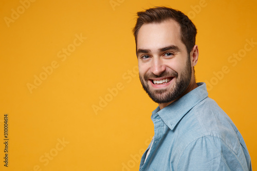 Side view of smiling young bearded man in casual blue shirt posing isolated on yellow orange background, studio portrait. People sincere emotions lifestyle concept. Mock up copy space. Looking camera. © ViDi Studio