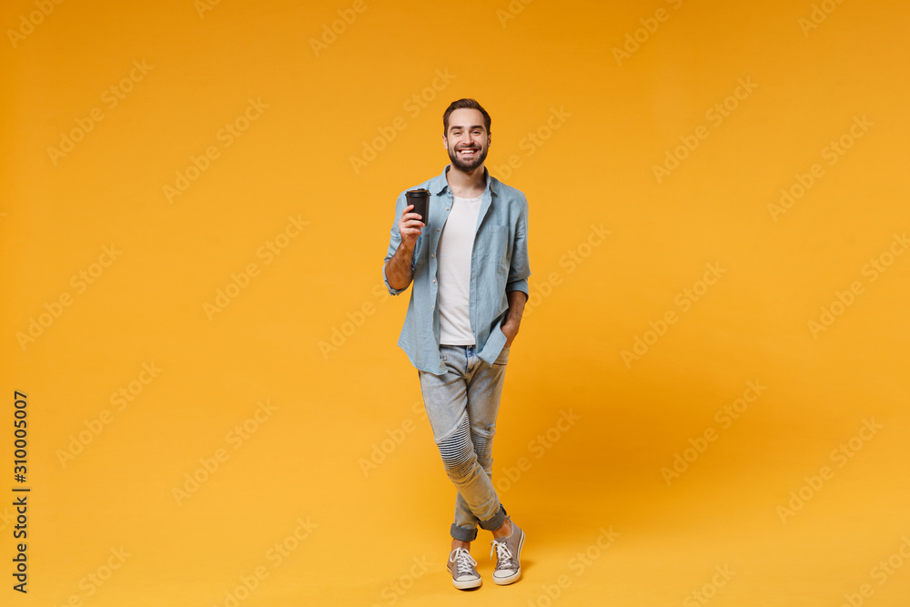 Fototapeta premium Smiling young man in casual blue shirt posing isolated on yellow orange wall background studio portrait. People sincere emotions lifestyle concept. Mock up copy space. Hold paper cup of coffee or tea.