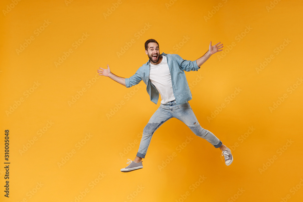 Excited young bearded man in casual blue shirt posing isolated on yellow orange wall background studio portrait. People emotions lifestyle concept. Mock up copy space. Jumping, spreading hands, legs.