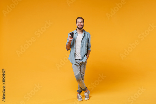 Smiling young man in casual blue shirt posing isolated on yellow orange wall background studio portrait. People sincere emotions lifestyle concept. Mock up copy space. Hold paper cup of coffee or tea.