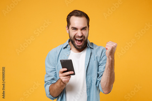 Happy young man in casual blue shirt posing isolated on yellow orange wall background studio portrait. People lifestyle concept. Mock up copy space. Hold mobile phone, doing winner gesture, screaming.