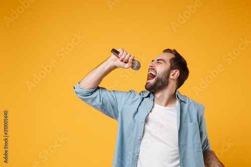 Handsome young bearded man in casual blue shirt posing isolated on yellow orange background, studio portrait. People sincere emotions lifestyle concept. Mock up copy space. Sing song in microphone.