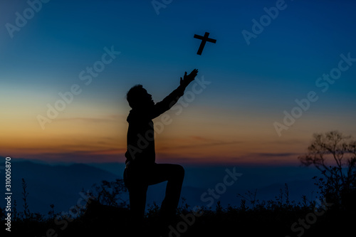 Silhouette of a young Christian man who is praying on the holy cell in the Bible and raising a cross over a Christian with a light sunset background. Christian concept