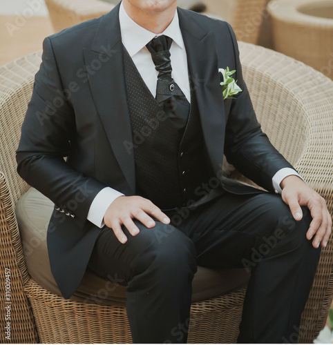 a young, fair-skinned man of 25 years, dressed in a beautiful elegant black suit with a white shirt and tie, is sitting in an armchair. Wedding image of the groom, fashion, style
