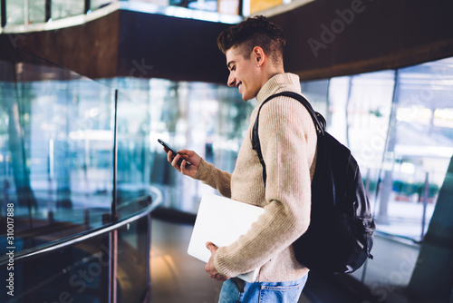 Smiling young man with backpack carrying laptop and using phone photo