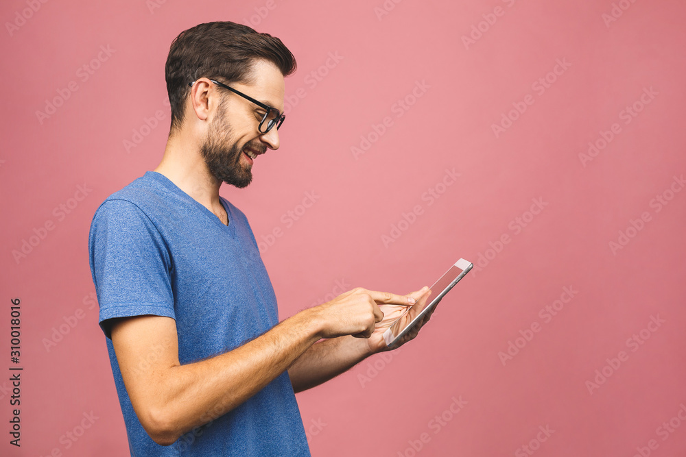 Happy young man in casual standing and using tablet isolated over pink background.