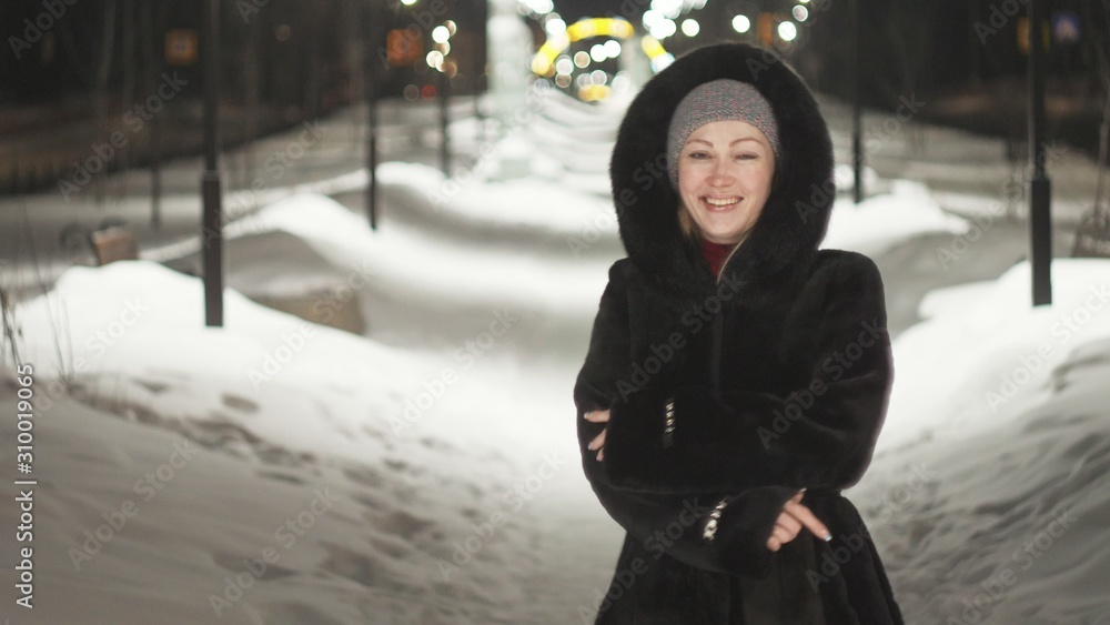 Pretty woman walking along the black fur coat. Outdoor. Girl walks on a winter night among the street lights of the city. Snow park with garlands. Beautiful sensual happy smile.