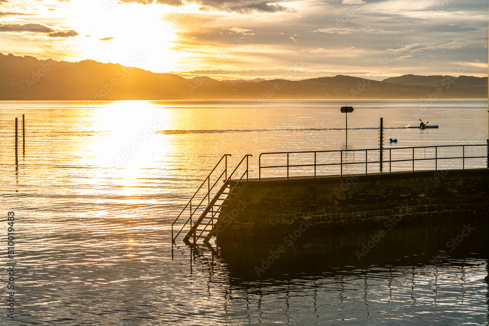 Obraz premium atmosphärische Abendstimmung (Sonnenuntergang) am Bodensee im Lindenhofpark mit Bergen, Wasserfläche und strahlendem Licht bei glaskarem HImmel (D, Bodensee, Lindau)