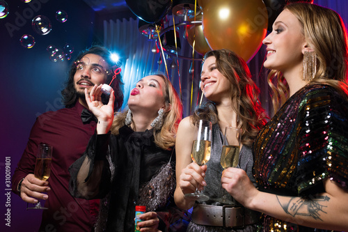 Glamorous girl blowing soap bubbles among friends with flutes of champagne