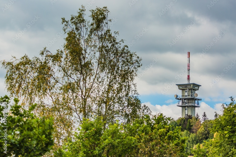 Obraz premium Zelena hora, Pelhrimov / Czech Republic - September 13 2019: Distant view of television communication tower with satellite dishes on. Cloudy sky. Green trees in foreground.