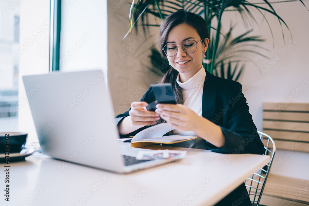 Positive businesswoman texting on smartphone while working on laptop at coffee shop Positive businesswoman texting on smartphone while working on laptop at coffee shop