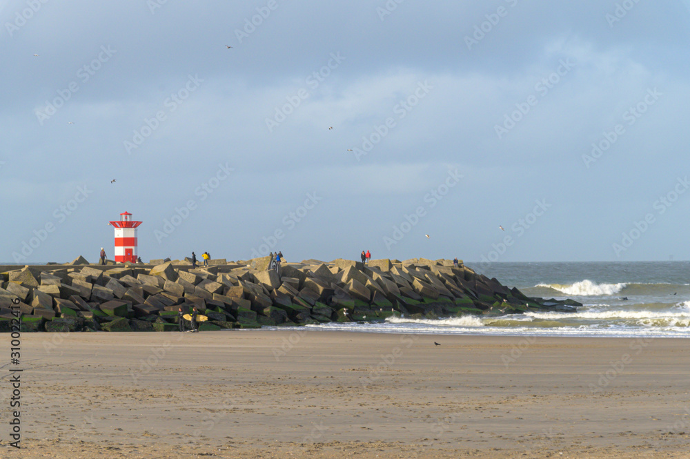 Breakwater of basalt blocks that have turned green  with a red-white beacon on the beach of Scheveningen.