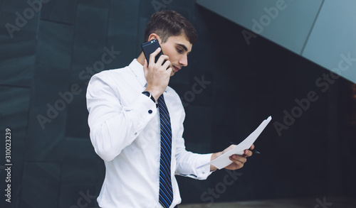 Confident young handsome businessman in formal outfit talking on mobile while cheeking papers in workplace