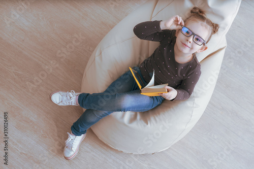 Schilderij op canvas Cute caucasian girl in glasses sitting on a bean bag and holding book