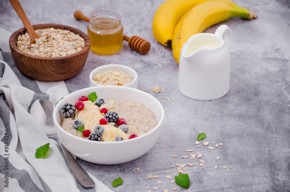 Oatmeal in a bowl with banana, frozen berries, almond petals, mint leaves and honey on a gray stone background. Tasty and healthy breakfast. Oatmeal in a bowl with banana, frozen berries, almond petals, mint leaves and honey on a gray stone background. Tasty and healthy breakfast.