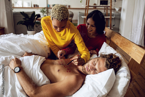 Young woman writing on chest of sleeping friend in bed at home