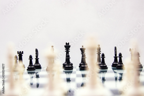 black chess pieces stand on a mirrored chess board with blurry white pieces in the foreground on a white background