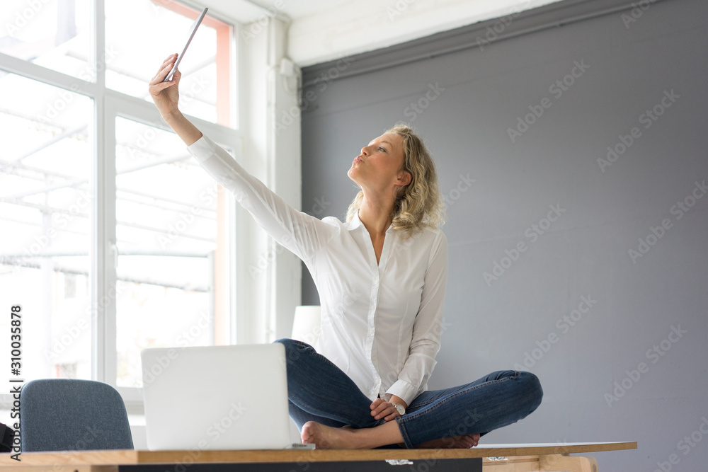 Young businesswoman sitting on desk in office taking a selfie with tablet Young businesswoman sitting on desk in office taking a selfie with tablet