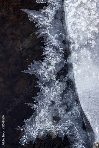 Fototapeta Naklejka Na Ścianę i Meble -  Ice on the river in winter. Picturesque hoarfrost, crystals on river ice over dark flowing water. Close-up. Selective focus. Vertical photo